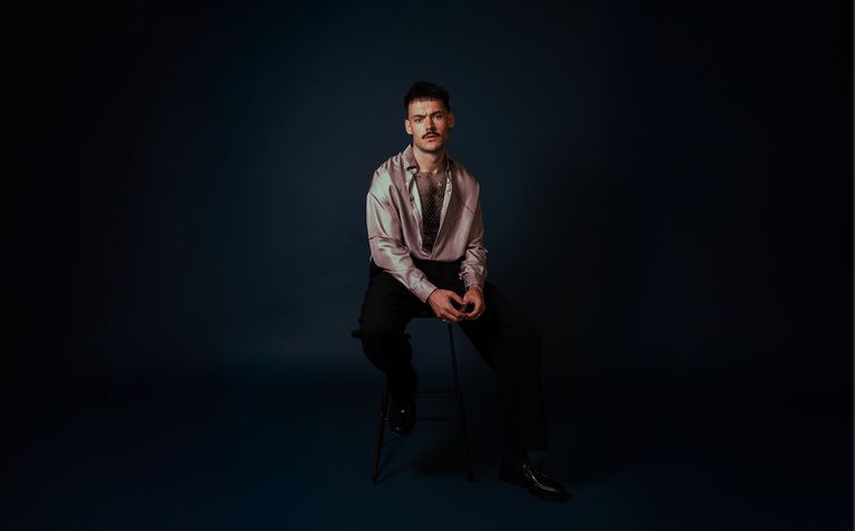 Musician James Marriott sitting on a stool in a satin shirt with a mustache against a dark blue studio background.